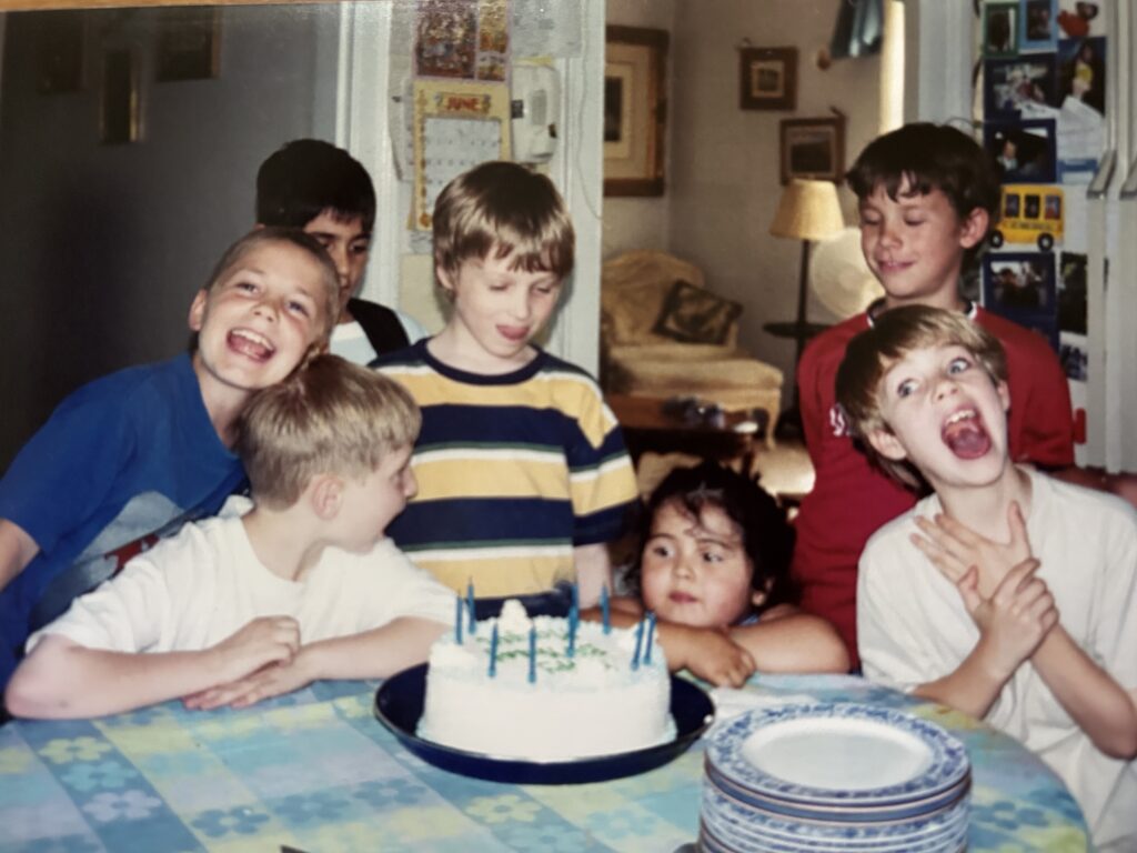 Redmond cheesing for the camera with friends at a table, clustered around a birthday cake, everyone smiling.